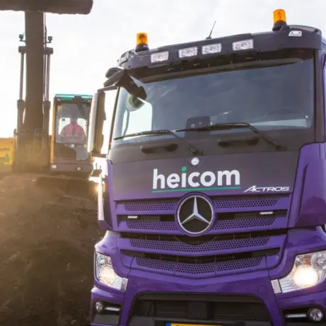 Paarse Heicom-vrachtwagen bij een gronddepot met graafmachine in tegenlicht