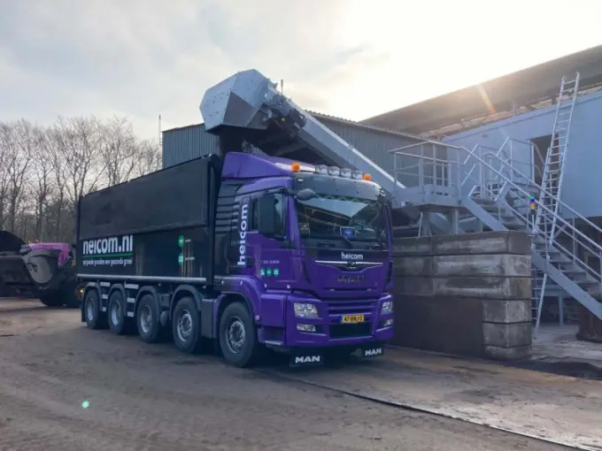 Paarse Heicom-vrachtwagen wordt via een transportband geladen bij een loods