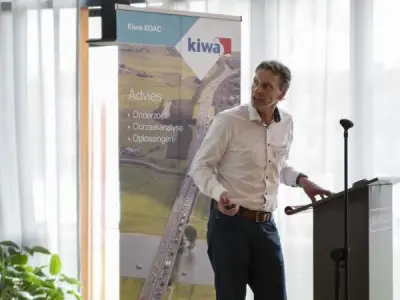 Spreker bij een katheder naast een roll-upbanner van Kiwa KOAC