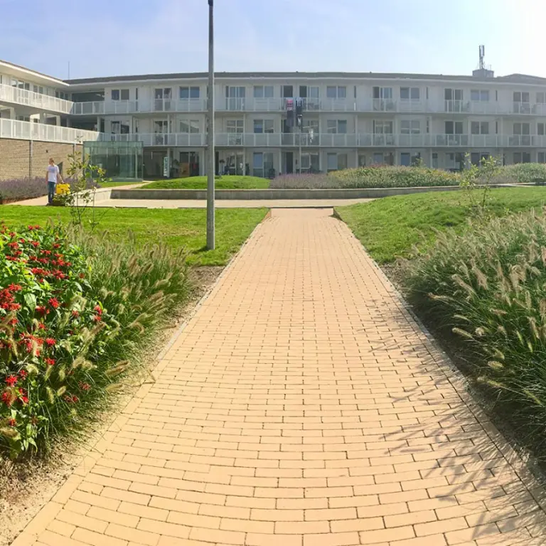 Klinkerpad tussen siergrassen en rode bloemen bij een appartementencomplex