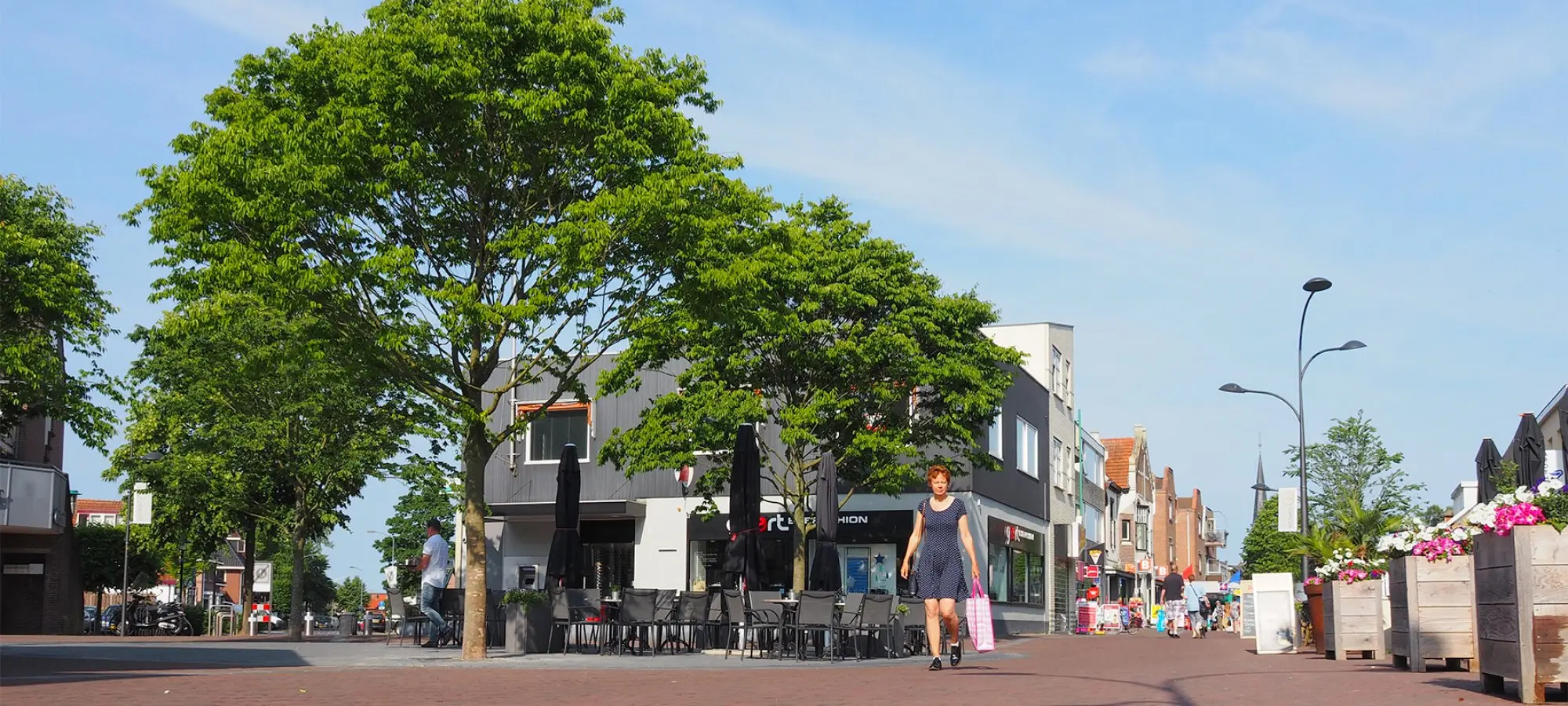 Zonnige winkelstraat met grote boom, terras en een vrouw met boodschappentas